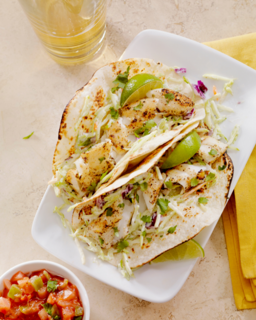 Two fish tacos with grilled tortillas on a white plate next to a bowl of salsa.