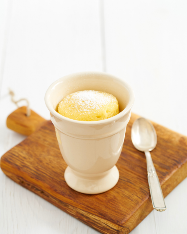 Vanilla Cake in a white mug sitting on a wooden cutting board with a spoon.