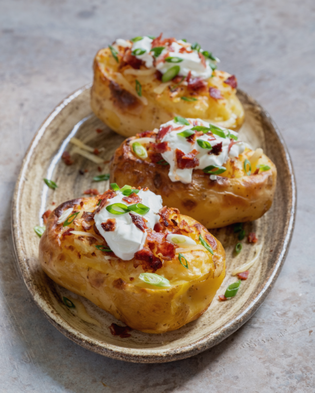 three loaded baked potatoes with sour cream, green onions, and bacon on a grey plate.