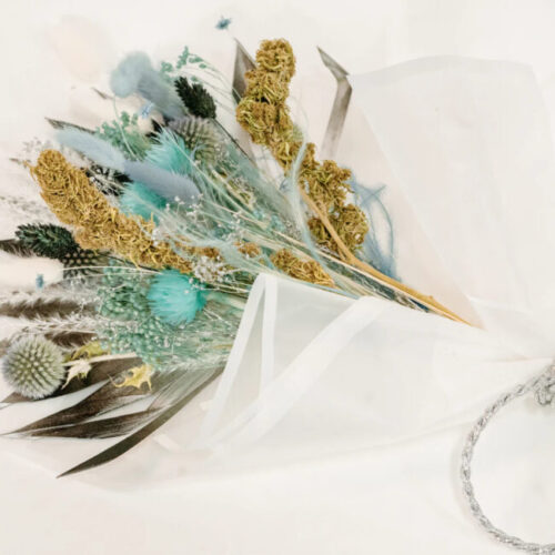 A delicate bouquet of dried CBD flowers and feathers in muted colors, nestled against white paper and ribbon.