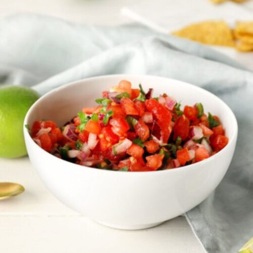 Pico De Gallo in a white small bowl with limes and a blue cloth in background