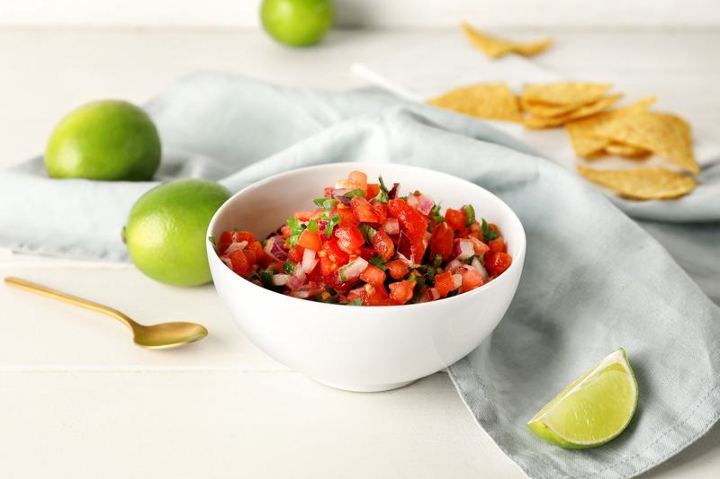 Pico De Gallo in a white bowl with limes, golden spoon and blue cloth